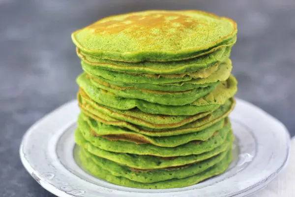 A rustic white plate holds a tall stack of bright green spinach pancakes.
