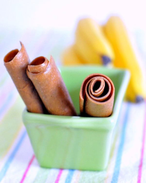 three peanut butter banana fruit roll (fruit leather made from pureed peanut butter and bananas) rolled up in tubes and sticking out of a small green ceramic loaf pan. a bunch of fresh bananas blurred in background, surface is a pastel striped napkin.