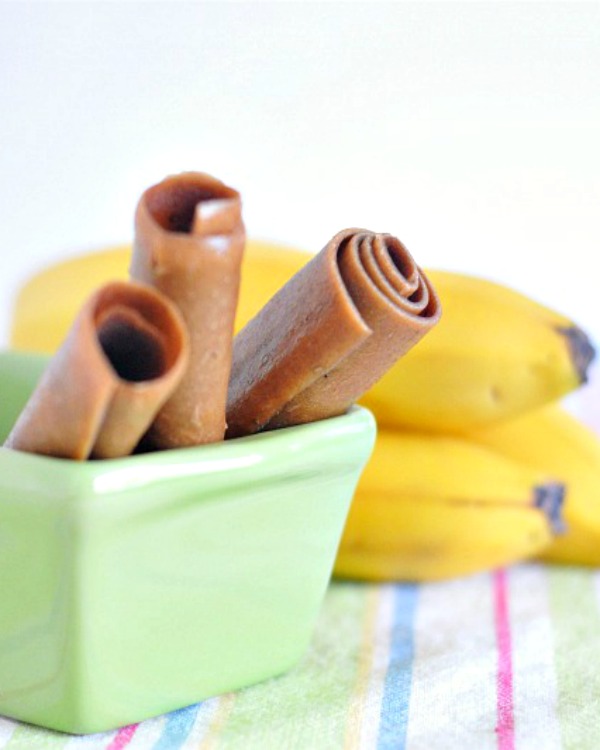 three peanut butter banana fruit roll (fruit leather made from pureed peanut butter and bananas) rolled up in tubes and sticking out of a small green ceramic loaf pan. a bunch of fresh bananas blurred in background, surface is a pastel striped napkin.