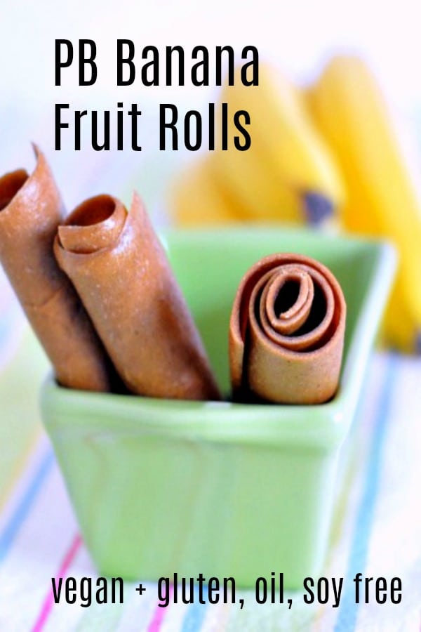 three peanut butter banana fruit roll (fruit leather made from pureed peanut butter and bananas) rolled up in tubes and sticking out of a small green ceramic loaf pan. a bunch of fresh bananas blurred in background, surface is a pastel striped napkin.