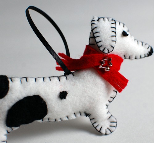 A handmade felt dachshund Christmas ornament. Dachshund is white with black spots and is wearing a red scarf.