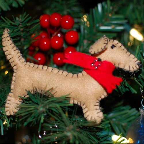 A handmade felt Christmas ornament sewn in the shape of a dachshund dog with a red scarf around the neck. Dachshund ornament is on a holiday tree with other ornaments and berries.