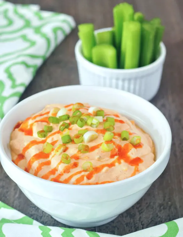 Vegan buffalo chicken dip in a white bowl, garnished with hot sauce and sliced green onion. set on a wood tabletop, with smaller bowls of celery slices for dipping.