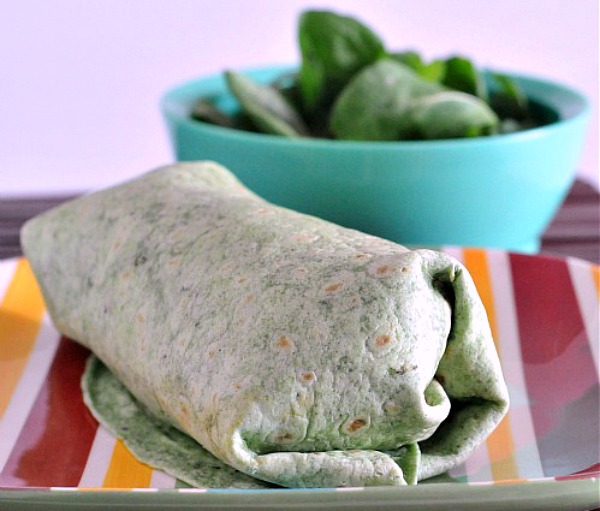 Chipotle Tofu Potato Burrito in a green spinach tortilla, sitting on a red and yellow striped plate, a light blue bowl of fresh spinach in background.