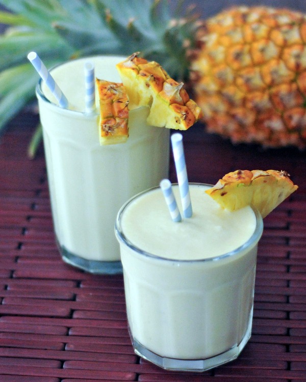 Lightened up pina coladas in glasses garnished with a wedge of pineapple and blue and white striped straws, with a whole fresh pineapple in background