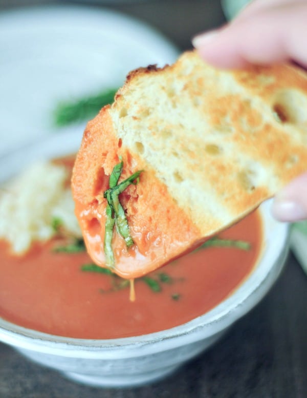 Tomato orange soup in a rustic white bowl, garnished with chopped basil and white rice, a slice of toasted bread being dipped in the bowl