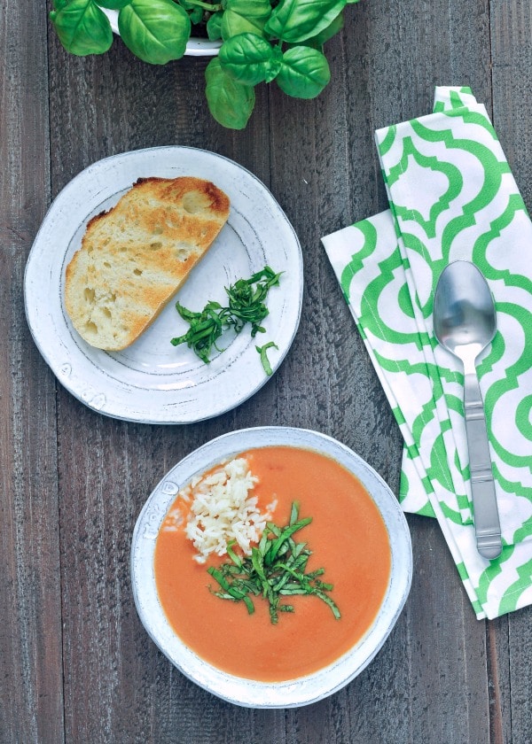 Tomato orange soup in a rustic white bowl, garnished with chopped basil and white rice, with a basil plant and a slice of crusty baguette on the side