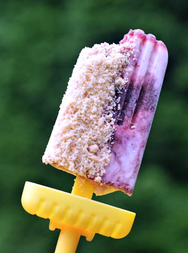 Cherry cheesecake popsicles with shortcake crumb on bright yellow handles against a green background of trees.