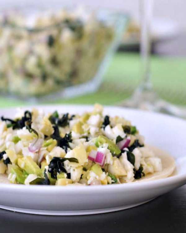 vegan artichoke shiitake ceviche with dark seaweed pieces and bright purple diced onion on a white dish. glass serving bowl of ceviche blurred in background.