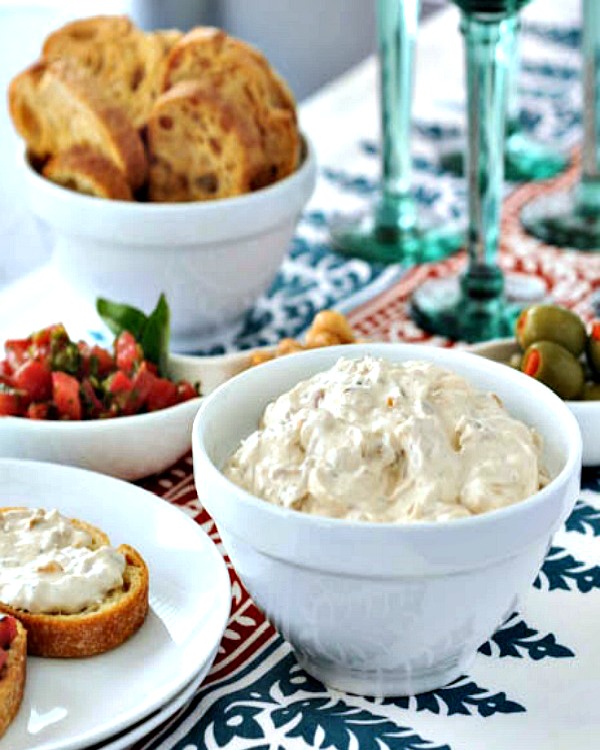a white serving bowl of caramelized shallot dip, with toasted baguette slices, a bowl of olives, and green stemmed wine glasses in background