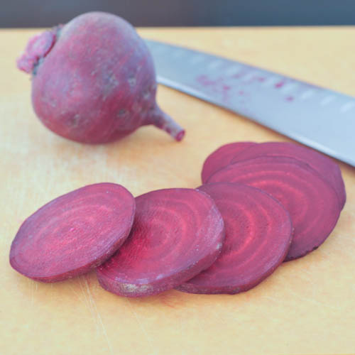 one whole beet and several slices of beet on a cutting board next to a knife.