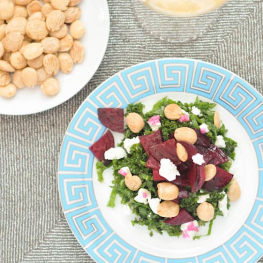 Roasted beet salad with vegan goat cheese, kale, and Marcona almonds on a plate with a teal and gold Greek key design on the rim. a small dish of Marcona almonds on the side.