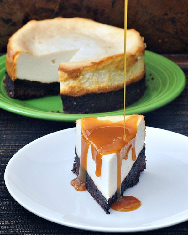 Brownie Bottomed Cheesecake slice on plate with caramel sauce poured over, whole cheesecake in background