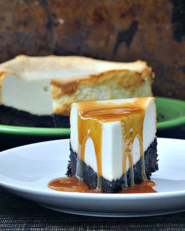 Brownie Bottomed Cheesecake slice on plate with caramel sauce poured over, whole cheesecake in background