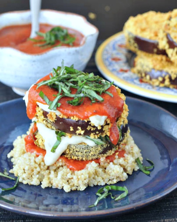 two slices of crispy eggplant stacked on top of quinoa and covered with marinara sauce and fresh chopped basil leaves. a bowl of marinara and a plate of crispy eggplant slices in background.