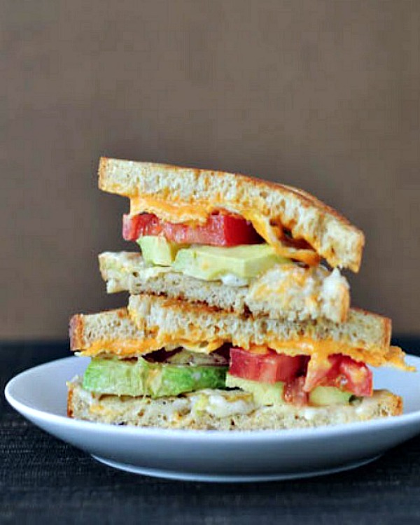 Salt and vinegar grilled cheese with avocado, tomato, potato chips against a brown background, stacked on a white plate