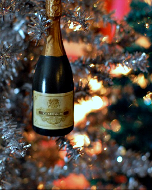 a small champagne bottle ornament hanging on a silver Christmas tree