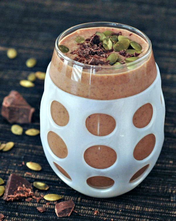 chocolate smoothie with in a glass with a white silicone sleeve, garnished with pepitas and shaved chocolate