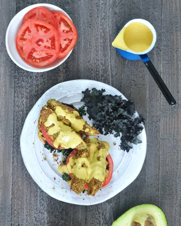 overhead view of Avocado Kale Florentine: fried avocado slices with tomato and kale on English muffins, on a white plate, tomato slices on the side