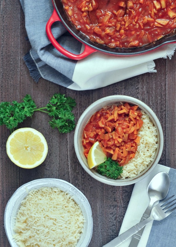 overhead view of red chicken and rice in a bowl with lemon slices and parsley garnish, on a white and grey napkin and dark wood background, half lemon and a fork on the side, with a larger bowl of rice and a skillet wit the remaining red chicken.