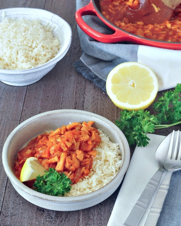 red chicken and rice in a bowl with lemon slices and parsley garnish, on a white and grey napkin and dark wood background, half lemon and a fork on the side, with a larger bowl of rice and a skillet wit the remaining red chicken.