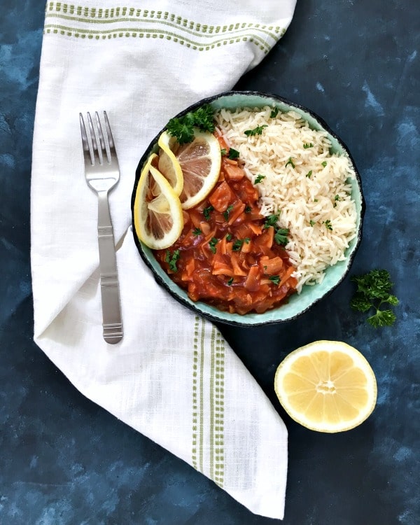 red chicken and rice in a bowl with lemon slices and parsley garnish, on a white napkin and dark blue background, half lemon and a fork on the side.