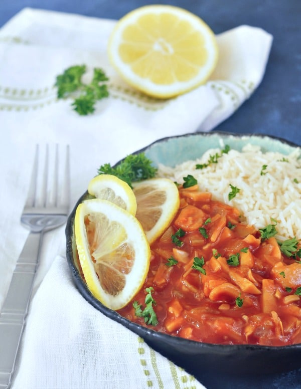 red chicken and rice in a bowl with lemon slices and parsley garnish, on a white napkin and dark blue background, half lemon and a fork on the side.