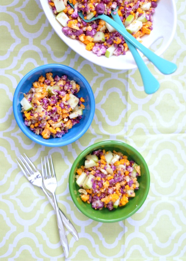 overhead view of cauliflower cashew confetti salad in a bright white serving bowl - this salad has purple, orange, and white cauliflower chopped small, with diced apple and a lemon sumac dressing