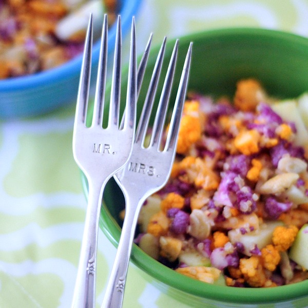 a "mr and mrs" fork set in front of a cauliflower cashew confetti salad in a green bowl - this salad has purple, orange, and white cauliflower chopped small, with diced apple and a lemon sumac dressing