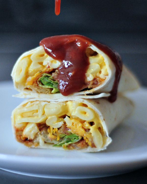 A BBQ mac and cheese burrito sliced in half on a white plate, with BBQ sauce dripping on top.