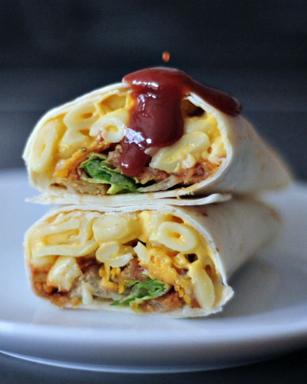 A BBQ mac and cheese burrito sliced in half on a white plate, with BBQ sauce dripping on top.