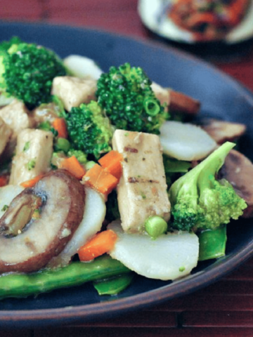 a dark blue plate of Moo Goo Gai Pan: sautéed vegan protein, sliced carrots, snap peas, broccoli trees over rice