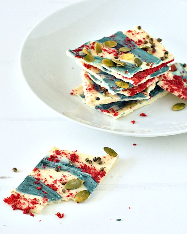 A stack of white chocolate bark on a white plate; bark is sprinkled with freeze dried raspberry powder, swirls of blue spirulina, and green pumpkin seeds.