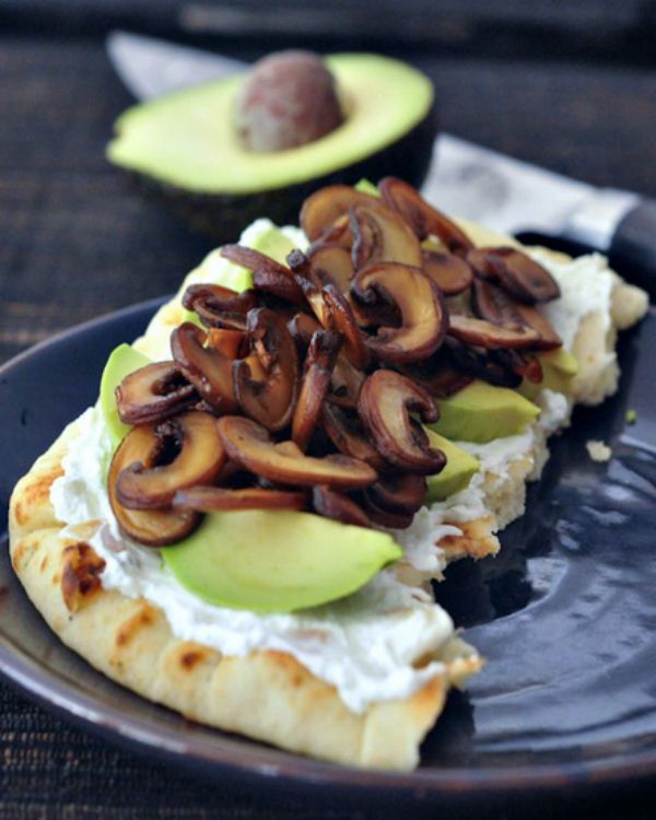 A slice of flatbread topped with vegan goat cheese, avocado slices, and sautéed mushrooms.