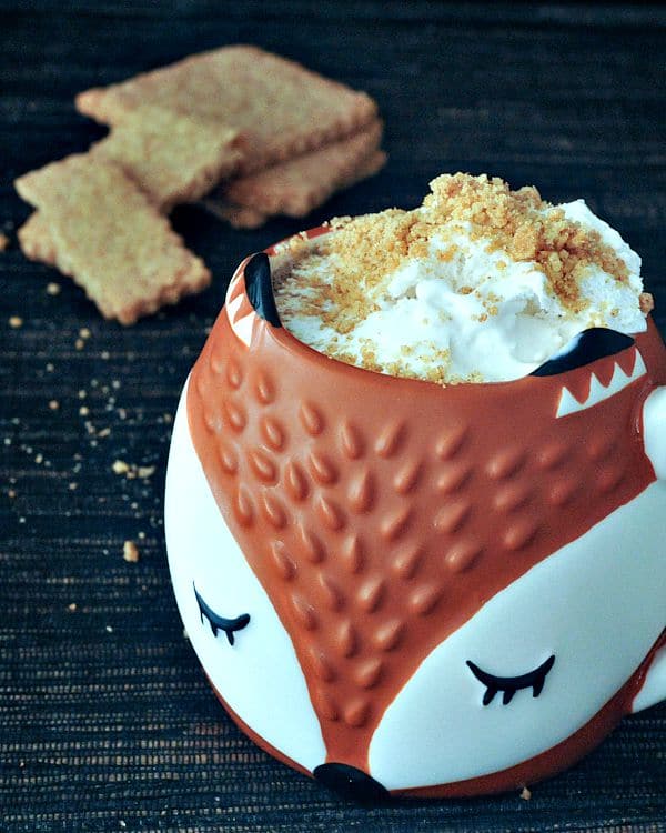 A fox face mug filled with a coffee latte topped with whipped cream and graham crackers.