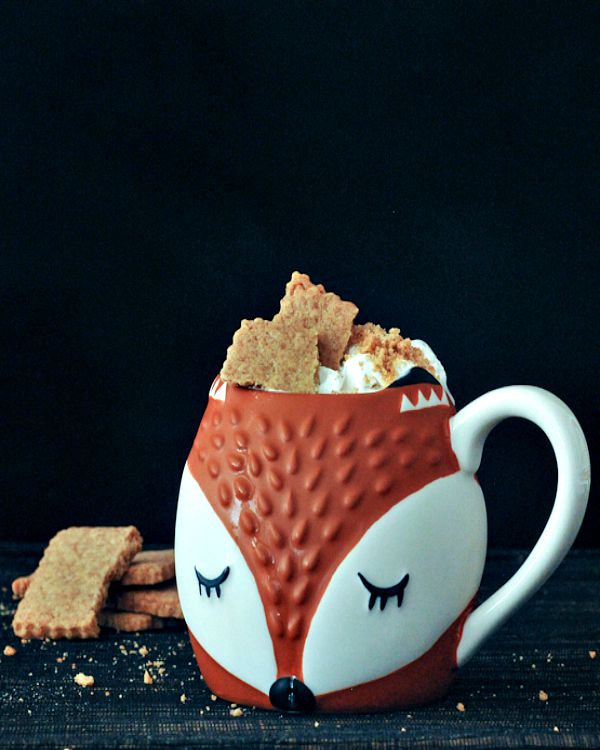 A fox face mug filled with a coffee latte topped with whipped cream and graham crackers.