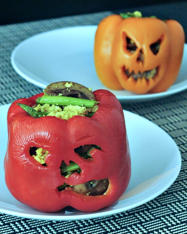 red and orange bell peppers carved with jack o lantern faces, cooked and filled with a breakfast mushroom scramble. on white plates against a black and white plaid background.