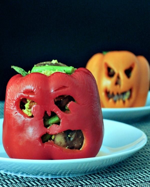 red and orange bell peppers carved with jack o lantern faces, cooked and filled with a breakfast mushroom scramble. on white plates against a black background.