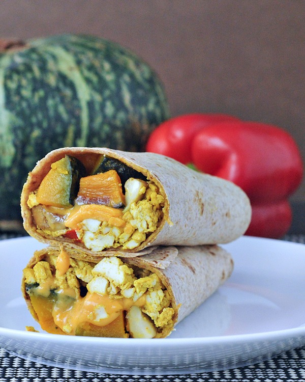 One cheesy kabocha breakfast burrito on a white plate, sliced in half to show filling (kabocha squash, vegan eggs, melty dairy free cheese).