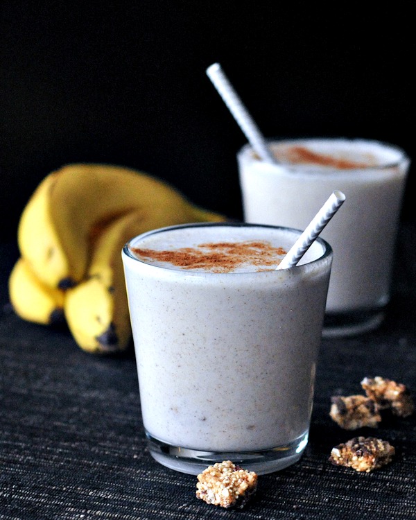 pale tan colored snickerdoodle smoothies in short glasses, garnished with cinnamon and granola, a bunch of banana in background