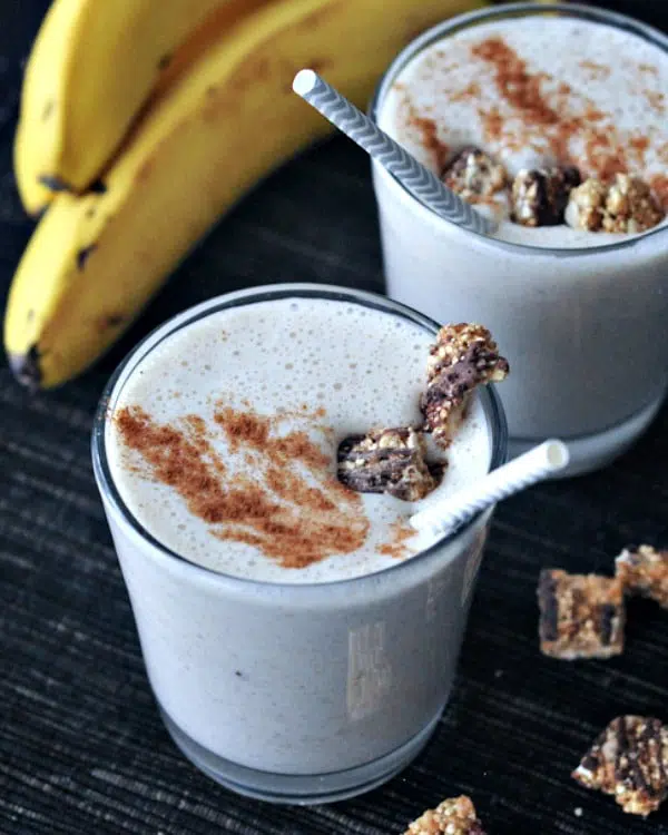 pale tan colored snickerdoodle smoothies in short glasses, garnished with cinnamon and granola, a bunch of banana in background