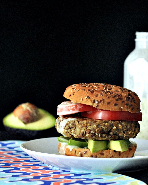 Sweet potato lentil burgers dressed with avocado slices, tomato slices, and a seeded bun on a white plate with a half avocado and ginger dressing bottle in dark black background