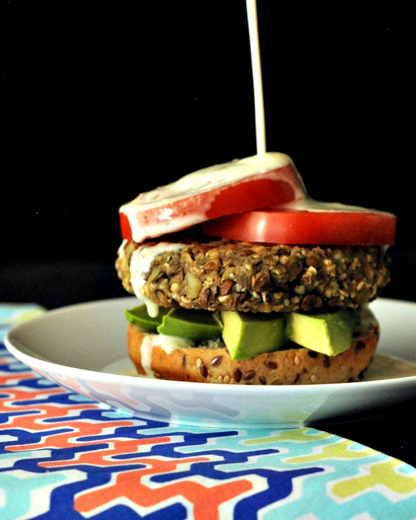 Sweet potato lentil burgers dressed with avocado slices, tomato slices, and a seeded bun on a white plate with ginger dressing bottle being poured over top