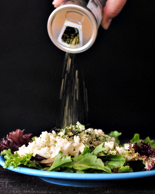 a hand shaking a jar of furikake flakes over a green salad with diced tofu, sriracha, sticky rice.
