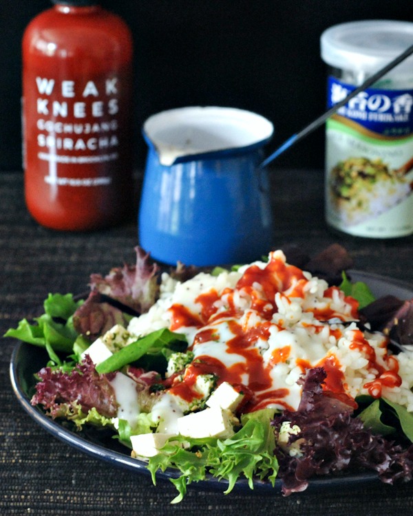 a green salad with diced tofu, sriracha, sticky rice and furikake flakes on a dark blue dish, drizzled with poppyseed dressing. a bottle of sriracha and jar of furikake in background.