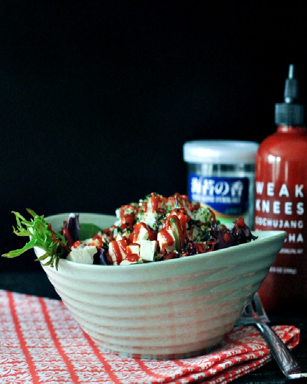 a green salad with diced tofu, sriracha, sticky rice, and furikake flakes in a light green bowl, drizzled with poppyseed dressing. a bottle of sriracha and jar of furikake in background.