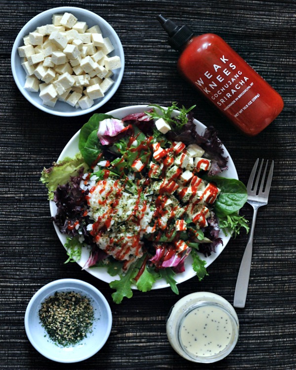 a green salad with diced tofu, sriracha, sticky rice, and furikake flakes on a white dish, drizzled with poppyseed dressing. a bottle of sriracha and jar of furikake in background.