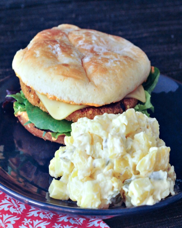 Classic vegan pickle potato salad on a dark blue plate next to a hamburger in a bun with lettuce and cheese.