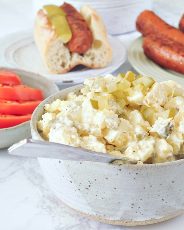 Classic vegan pickle potato salad in a rustic grey serving bowl, hot dogs in buns and sliced tomatoes on plates in background