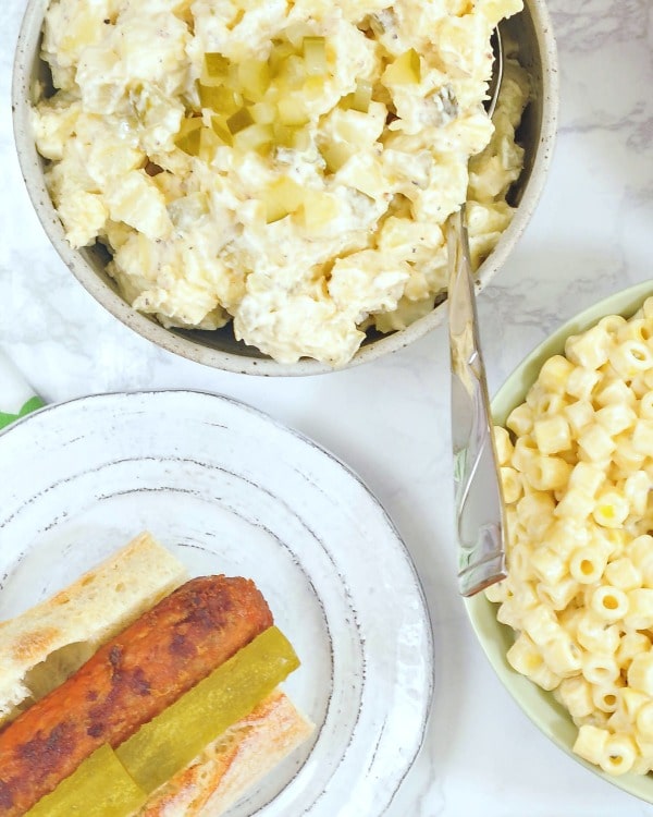 overhead view of classic vegan pickle potato salad in a bowl, next to a bowl of macaroni salad and a plate with a hot dog in a bun